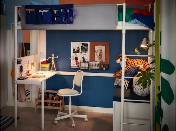 Ei hvit bordlampe lyser opp et skrivebord og en blå arbeidsstol for barn under ei loftseng.