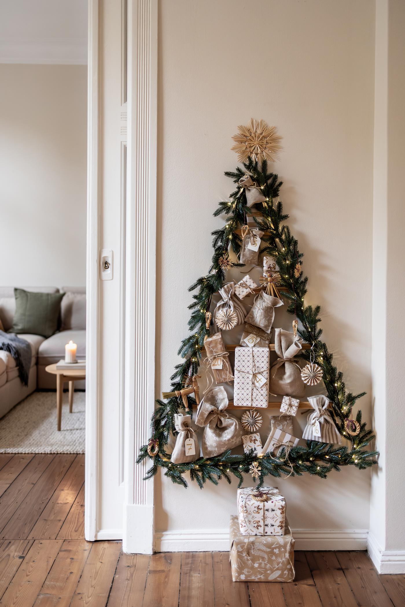 Ein Adventskalender im natürlichen Look wurde mit Kindern gebastelt. Ein Tannenbaum hängt mit bunten Kugeln an einer weißen Wand.