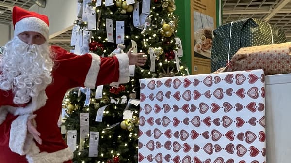 Ein als Weihnachtsmann verkleideter Mann steht vor einem festlich geschmückten Weihnachtsbaum mit goldenen Kugeln und Wunschzetteln. Er zeigt auf große, eingepackte Geschenke mit weihnachtlichem Muster.  