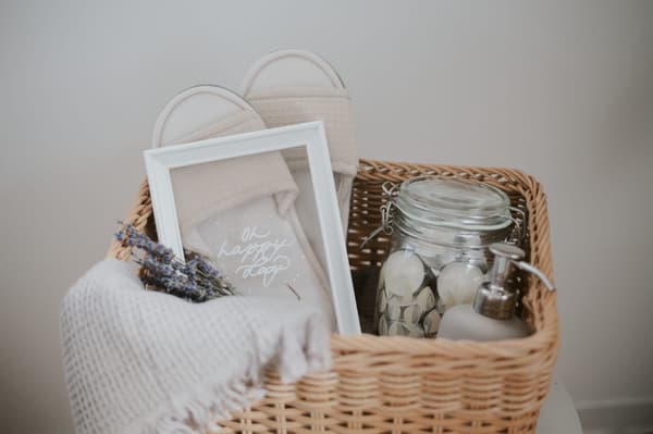 Ein aus Naturfasern geflochtener Geschenkkorb BEKNA, gefüllt mit einem weißen Bilderrahmen mit Gruß, Teelichtern in einem KORKEN Einmachglas, einem Seifenspender und beigne SKOVELSJÖN Pantoffeln. 