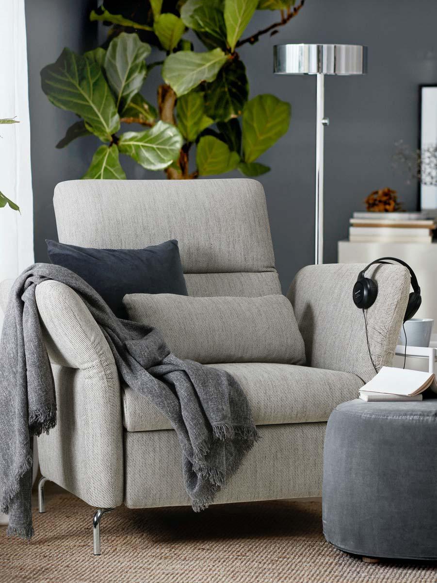 Ein beigefarbener FAMMARP Sessel mit verstellbaren Armlehnen und Rückenlehne steht vor einer grauen Wand. Hinter dem Sessel steht eine hohe Zimmerpflanze und eine silberfarbene Stehleuchte.