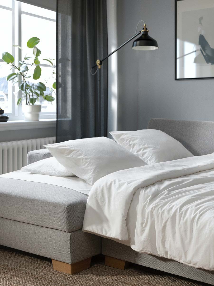 Ein beigefarbenes SÖRVALLEN Bettsofa mit Recamiere steht aufgeklappt in einem Wohnzimmer. Darauf liegt weißes Bettzeug.