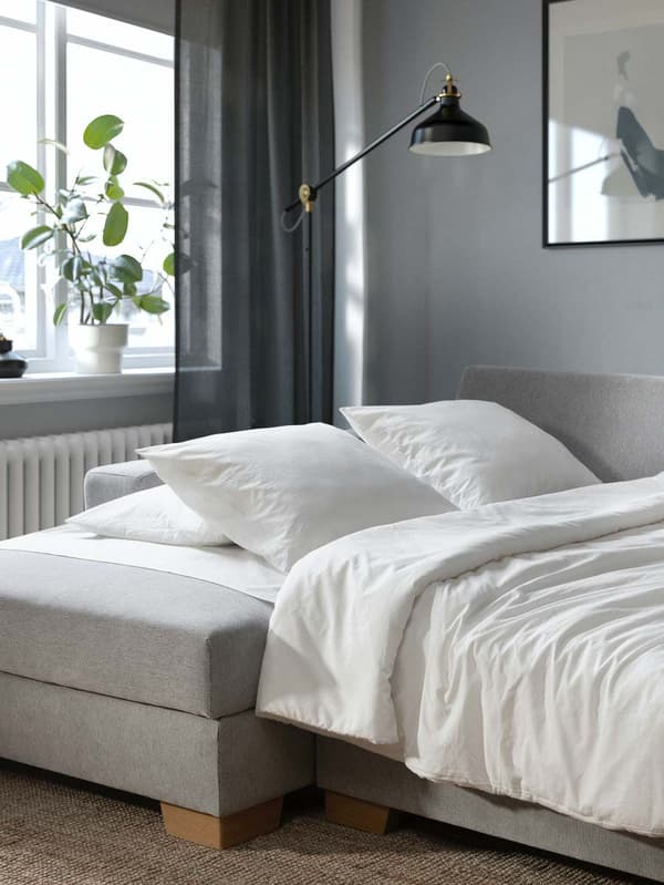 Ein beigefarbenes SÖRVALLEN Bettsofa mit Recamiere steht aufgeklappt in einem Wohnzimmer. Darauf liegt weißes Bettzeug. 