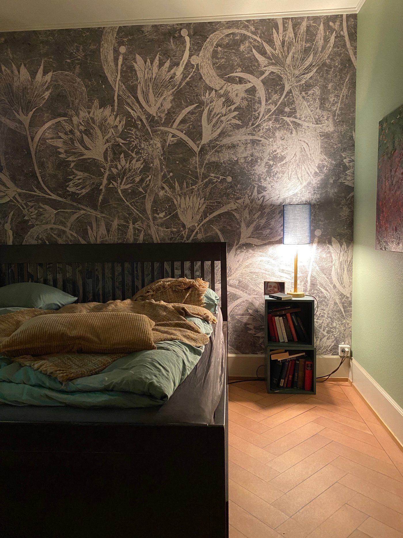 Ein Bett mit Bettwäsche darauf steht an einer Wand mit Fototapete mit floralem Muster. Daneben ein kleiner Nachttisch mit Büchern darin und einer Nachttischlampe darauf.