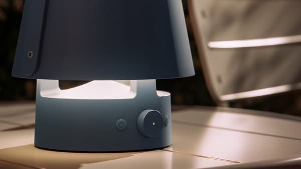 Ein blauer VAPPEBY Bluetooth-Lautsprecher steht am Abend auf einem Gartentisch. Die Leuchte ist eingeschaltet.