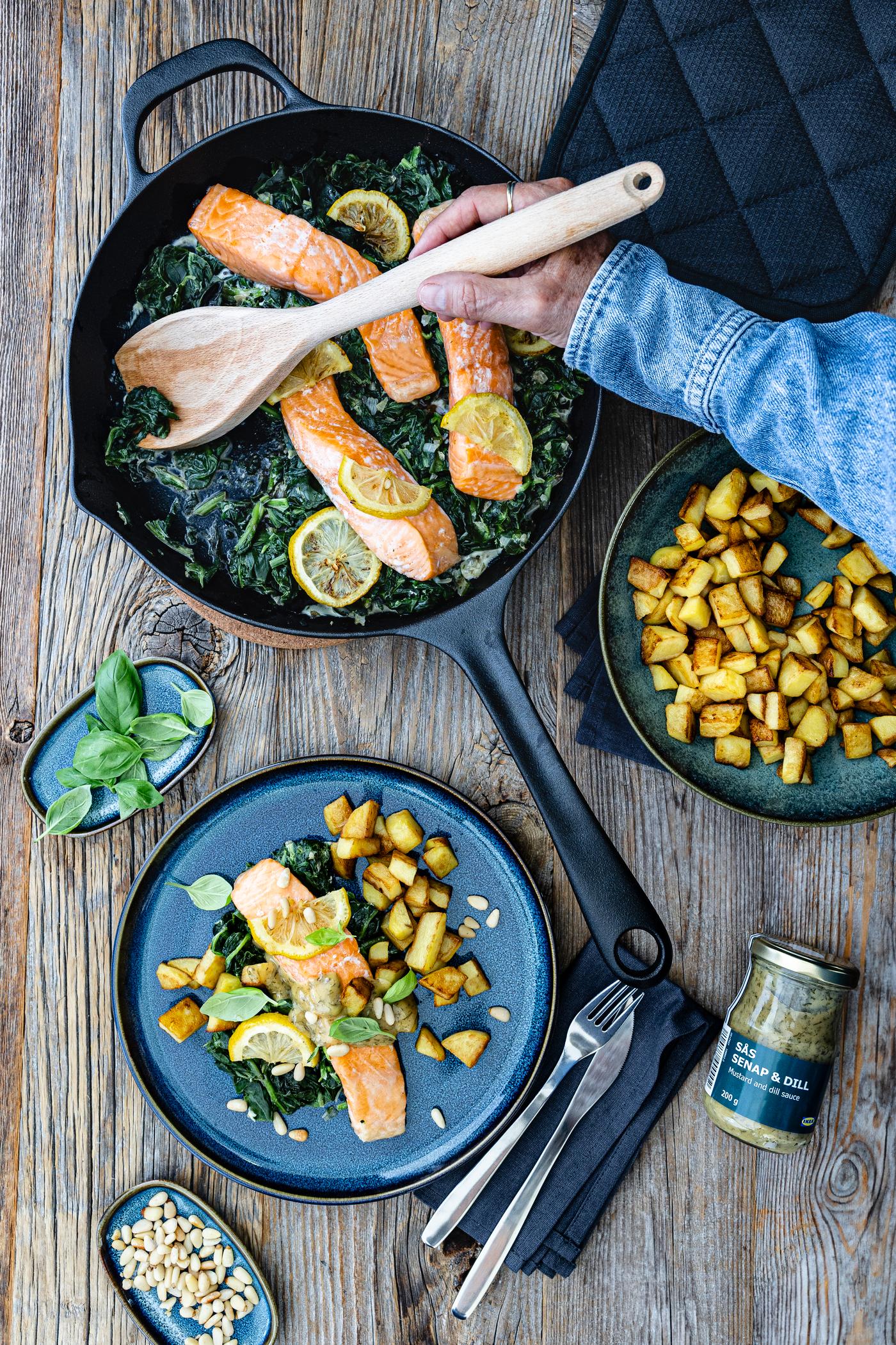 Ein Blick von oben in die Pfanne mit Spinat, Lachsfilet und Zitronenscheiben. Daneben stehen 2 Teller, die mit dem fertigen Gericht befüllt sind.