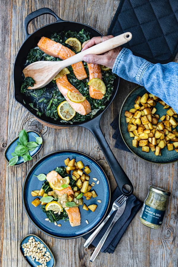 Ein Blick von oben in die Pfanne mit Spinat, Lachsfilet und Zitronenscheiben. Daneben stehen 2 Teller, die mit dem fertigen Gericht befüllt sind.
