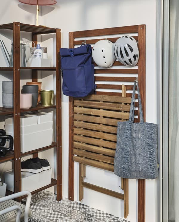 Ein braun lasiertes ÄPPLARÖ Wandpaneel an einer Wand. An Haken sind dort ein Rucksack, eine Tasche, ein Klapptisch und zwei Fahrradhelme zu sehen.