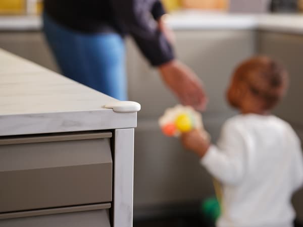 Ein erwachsener Mensch und ein Kleinkind sind im Hintergrund des Bildes in einer Küche zu sehen. An einer Arbeitsplatte im Vordergrund ist ein UNDVIKA Eckschutz befestigt.