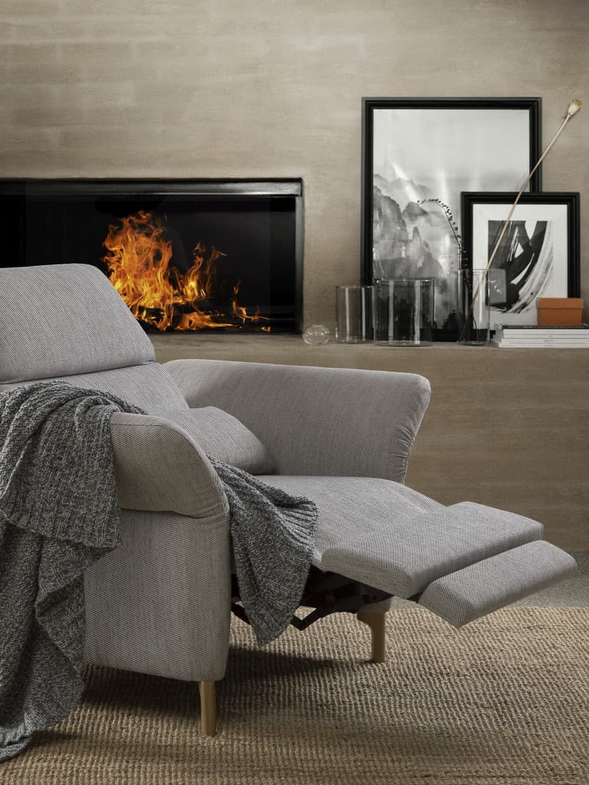 Ein FAMMARP Relaxsessel mit elektrisch verstellbarem Fußteil und verstellbaren Armlehnen und Rückenlehnen steht vor einem Kamin in dem ein Feuer brennt.