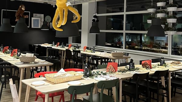 Ein festlich gedeckter Essbereich in einem IKEA-Restaurant mit langen Holztischen und schwarzen Stühlen. Auf den Tischen stehen Teller, Körbe mit Brot, Laternen mit Kerzen und rote Servietten. An den schwarzen Deckenlampen hängen Kuscheltiere, darunter ein großer gelber Oktopus und ein Affe.  