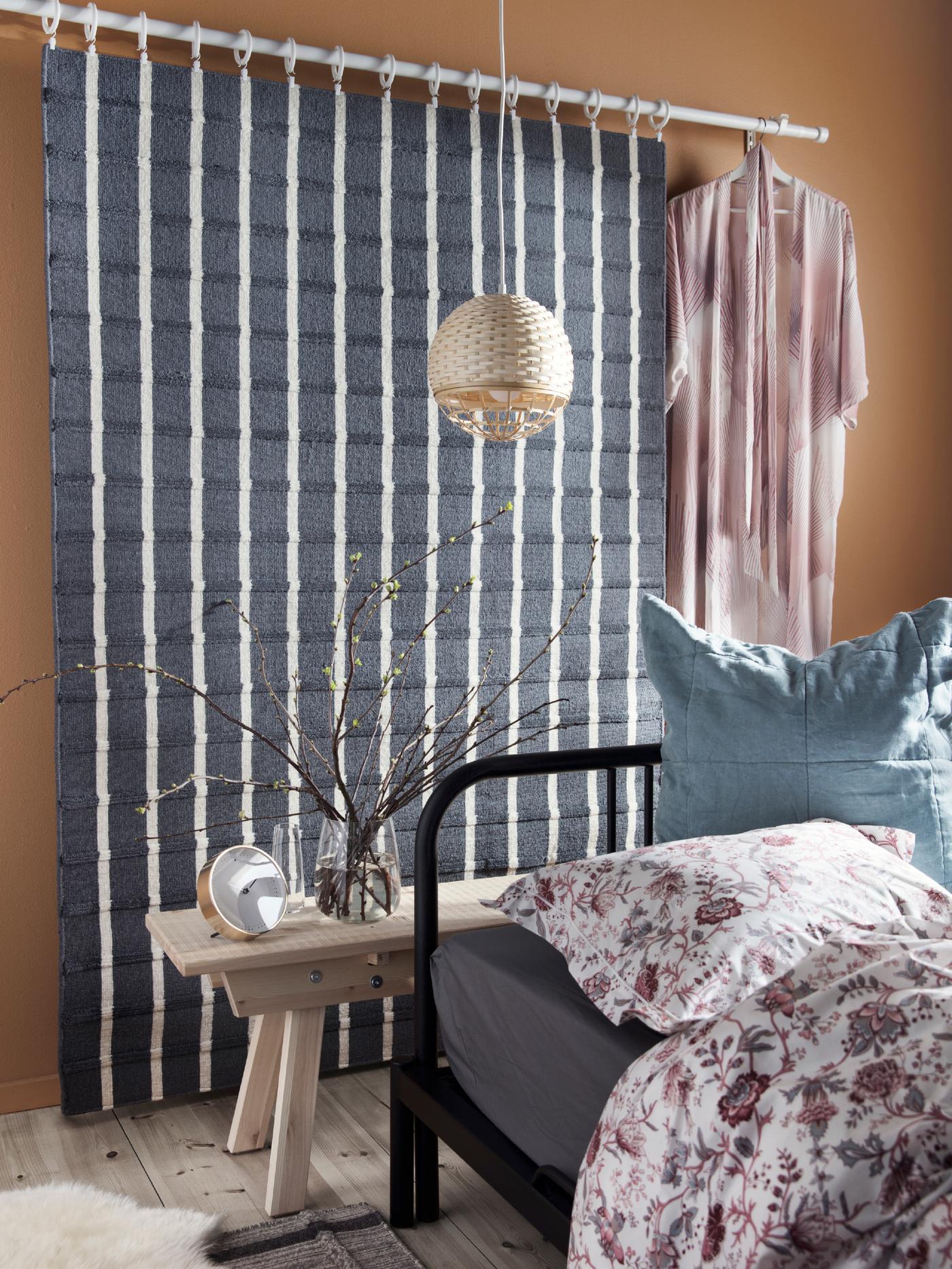 Ein FOULUM Teppich in Grau und Weiß und ein Morgenmantel an einer Gardinenstange hinter einem Bett.