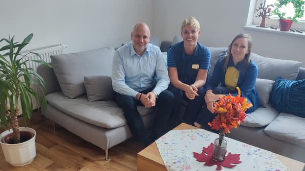 Ein fröhlicher Mann mit hellblauem Hemd und zwei lächelnde Frauen in IKEA Bekleidung sitzen auf einem grauen Ecksofa. Links neben dem Sofa steht eine Pflanze, vor dem Sofa steht ein Beistelltisch. Auf diesem steht ein roter Herbststrauß in einer Vase. 