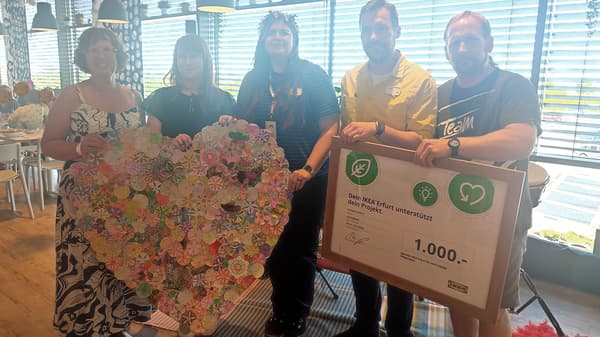 Ein fröhlicher Mann und ein IKEA Mitarbeiter halten einen Spendencheck in Höhe von 1.000.- Euro in den Händen. Neben ihnen stehen zwei fröhliche Frauen und eine IKEA Mitarbeiterin und halten ein großes Herz, welches mit Papierblumen beklebt ist.