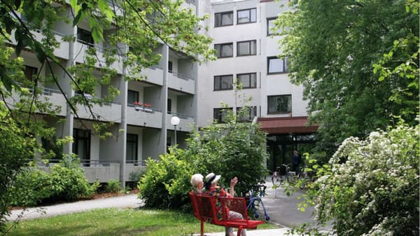 Ein Gartenbereich vor einem mehrstöckigen Wohngebäude mit Balkonen. Zwei ältere Personen sitzen auf einer roten Bank und genießen die grüne Umgebung. Büsche, Bäume und blühende Pflanzen umgeben den Sitzbereich, während im Hintergrund Fahrräder und ein Eingang zu sehen sind.  