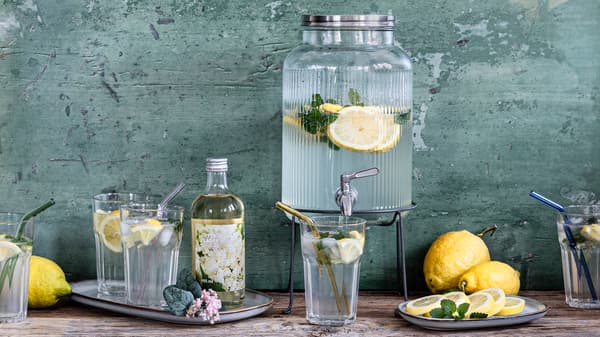 Ein Gefäß mit Zapfhahn, gefüllt mit selbstgemachter Holunder-Zitronenlimonade. Daneben befinden sich frische Zutaten und Gläser.