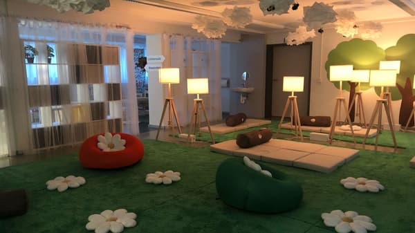 Ein gemütlicher Innenraum, gestaltet wie ein Garten: weiße Blumenkissen, orange und grüne Sitzsäcke, wolkenartige Deckenleuchten, Wanddekorationen mit Bäumen und ein kleiner Waschbereich hinter transparenten Vorhängen.