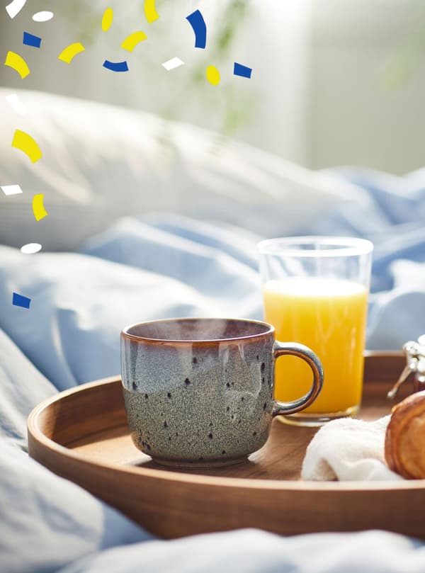 Ein GLADELIG Becher in beigegrün, ein Glas Orangensaft und ein Croissant stehen auf einem Tablett in einem Bett.