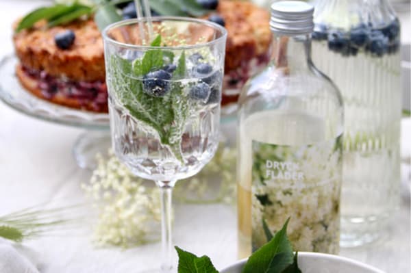 Ein Glas Limonade mit Blaubeeren und Minze auf einem Tisch. Im Hintergrund steht ein Blaubeerkuchen.
