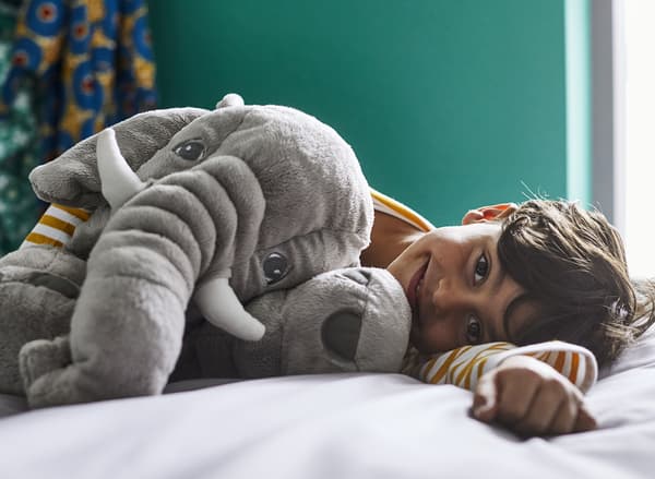 Ein glücklich lächelndes Kind liegt auf einem Bett in einem dunkelgrünen Zimmer und hält ein JÄTTESTOR Stoffspielzeug in Elefantenform im Arm.
