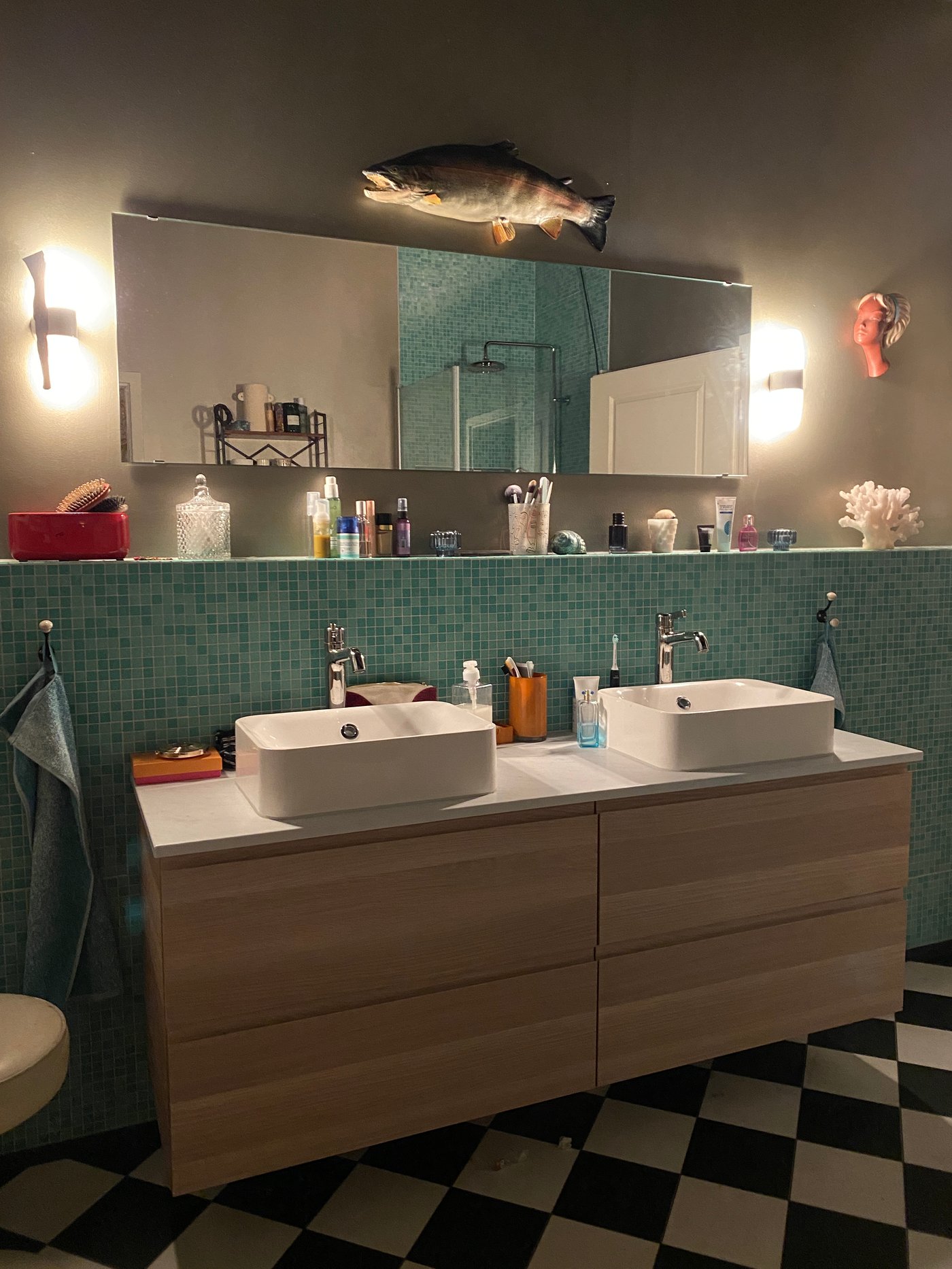 Ein GODMORGON Doppelwaschtisch mit Schubladen und einem grossen, beleuchtetem Spiegel darüber. Auf der Fläche unter dem Spiegel und neben den Waschbecken stehn viele Badutensilien.