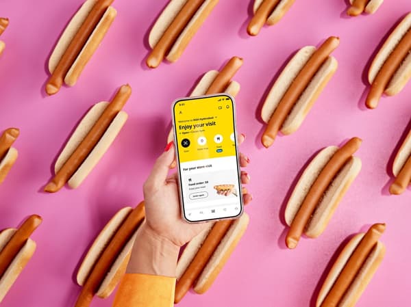 Ein grafischer Hintergrund mit mehreren IKEA Hotdogs; im Vordergrund hält eine Hand ein Smartphone, auf dem eine Essensbestellung in der IKEA App angezeigt wird.