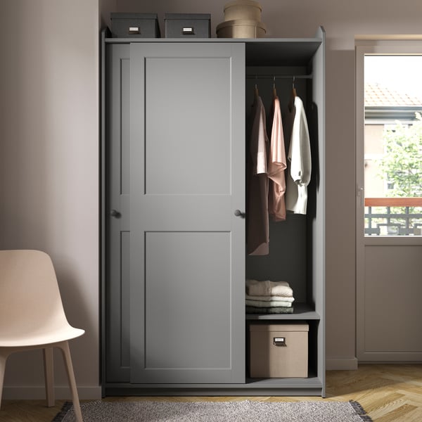 Ein grauer HAUGA Kleiderschrank mit Schiebetüren steht neben einer Tür. Sein Inhalt ist zu sehen, der aus Kleidung an Bügeln und Boxen besteht.