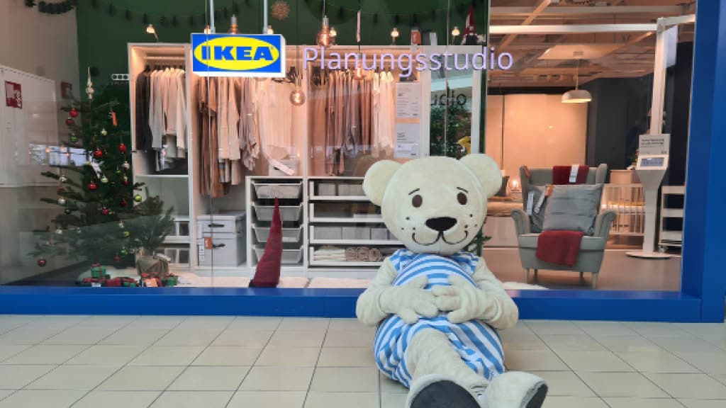 Ein Grauer Tedibär in einem Blau Weißem Streifenanzug sitzt andgeleht gegen die Front eines Ikea Planungstudios 