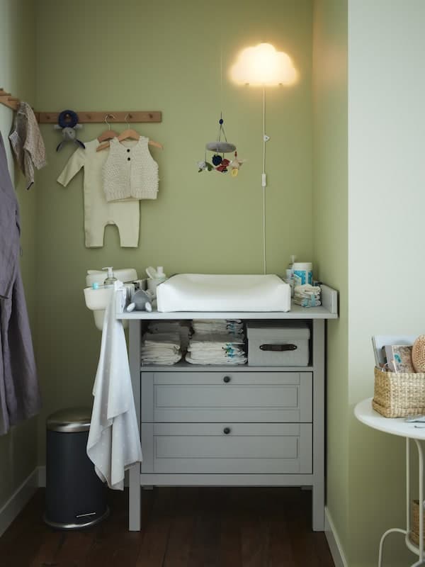 Ein grauer Wickeltisch/-kommode, Hakenleisten aus Eiche mit Babykleidung, einer Wickelunterlage und einer LED-Wandleuchte in Form einer Wolke.