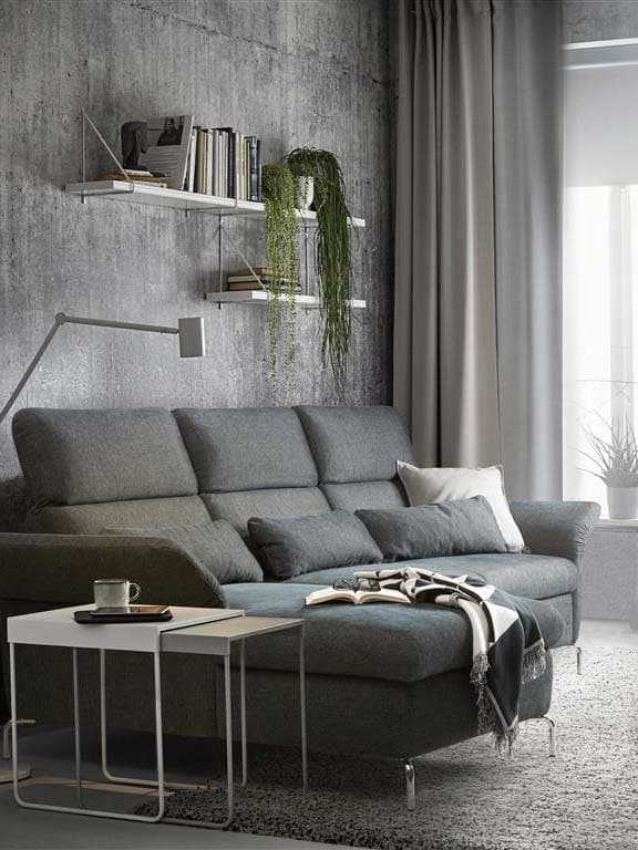 Ein graues FAMMARP 3er Sofa mit Recamiere und verstellbaren Rücklehnen und Armlehnen steht vor einer grauen Betonwand. 