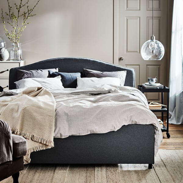 Ein graues HAUGA Doppelbett mit heller Bettwäsche steht in einem sonnenlichtdurchfluteten Schlafzimmer, neben dem Bett befindet sich ein Ablagetisch.