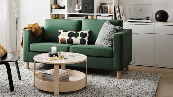 Ein grünes PÄRUP Sofa mit zwei gemusterten Kissen in einem weiß gehaltenen Wohnzimmer mit offenen Regalen.