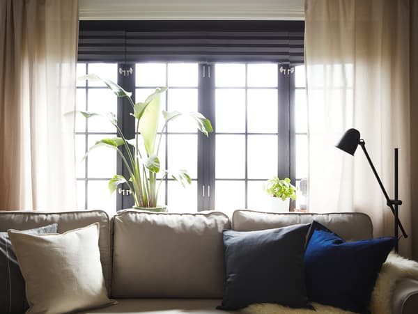 Ein hellbeiges Bettsofa steht vor einer Fensterfront mit beigefarbenen Gardinen und grauen Verdunklungsrollos.