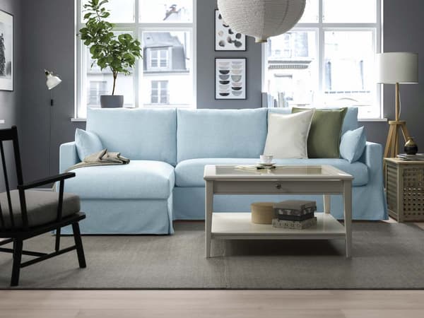 Ein hellblaues Eckbettsofa steht vor 2 Wohnzimmerfenstern. Davor liegt ein Teppich auf dem ein rechteckiger weißer Couchtisch steht. Der Couchtisch hat eine Glasplatte und eine Schublade. 