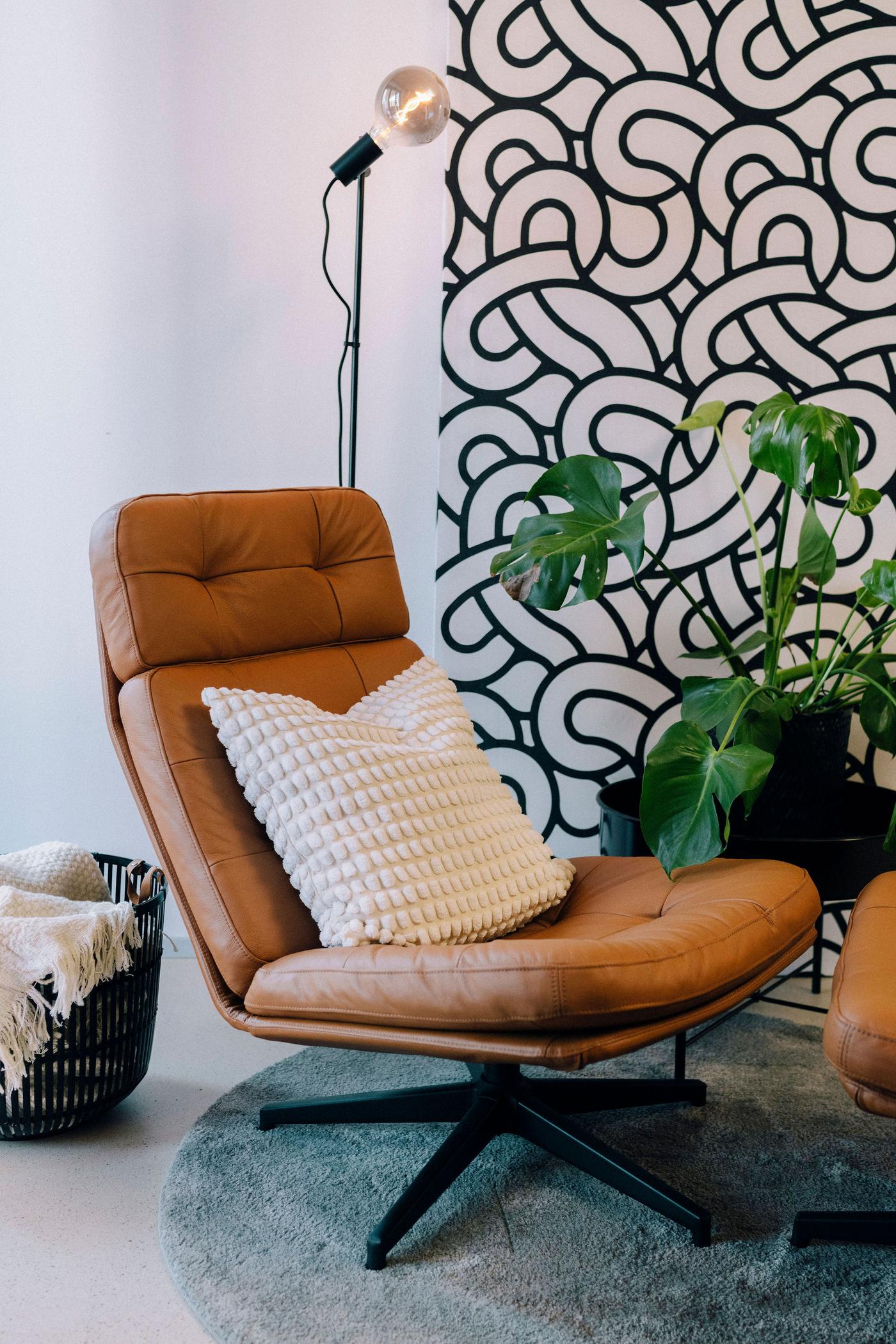 Ein hellbrauner, moderner Ledersessel mit einem Kissen steht neben einer Stehlampe und einer Pflanze vor einer Wand.