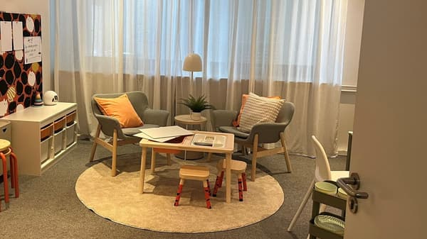 Ein heller, gemütlicher Raum mit zwei grauen Sesseln mit orange-weißen Kissen, weißer Stehlampe und Pflanze auf dem Tisch. Links ein weißes Regal mit bunten Dekorationen, im Hintergrund große Fenster mit transparenten Vorhängen.