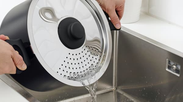 Ein HEMLAGAD Topf mit Deckel mit Ausguss wird über einer Edelstahlspüle gehalten um das Wasser abzulassen.