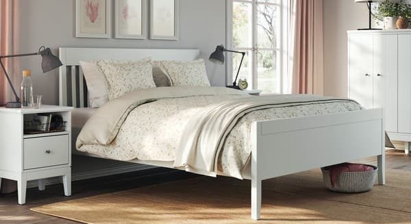 Ein IDANÄS Bettgestell, ein Ablagetisch und zwei Kommoden in Weiß stehen in einem Schlafzimmer; auf dem Bett liegt Bettzeug in LUKTJASMIN Bettwäsche-Set in Dunkelgrau.