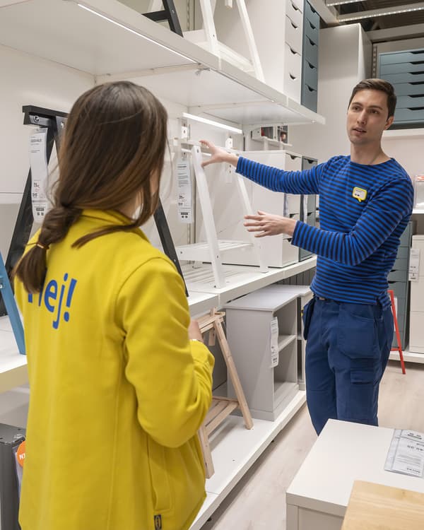 Ein IKEA-Mitarbeiter in blauem Streifenshirt erklärt einer Kollegin in gelber „Hej!“-Jacke verschiedene Tischböcke in einer Regalreihe der Ausstellung.