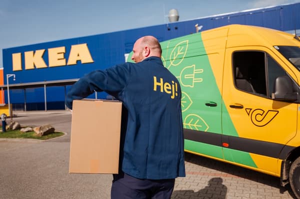 Ein IKEA Mitarbeiter trägt ein Paket vor einem IKEA Einrichtungshaus und einer Post Lieferwagen. 