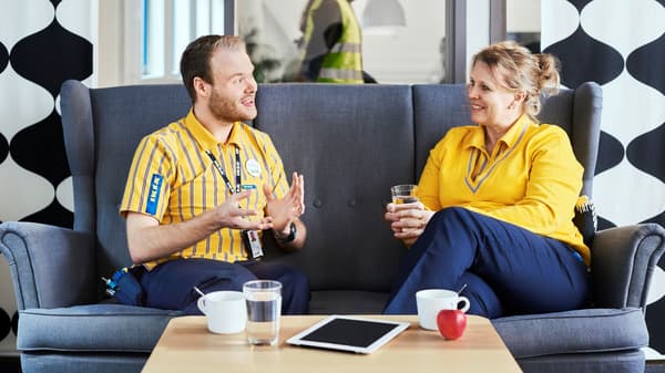 Ein IKEA Mitarbeiter und eine IKEA Mitarbeiterin sitzen auf einem grauen Sofa und unterhalten sich.