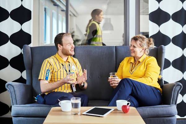 Ein IKEA Mitarbeiter und eine IKEA Mitarbeiterin sitzen auf einem grauen Sofa und unterhalten sich.