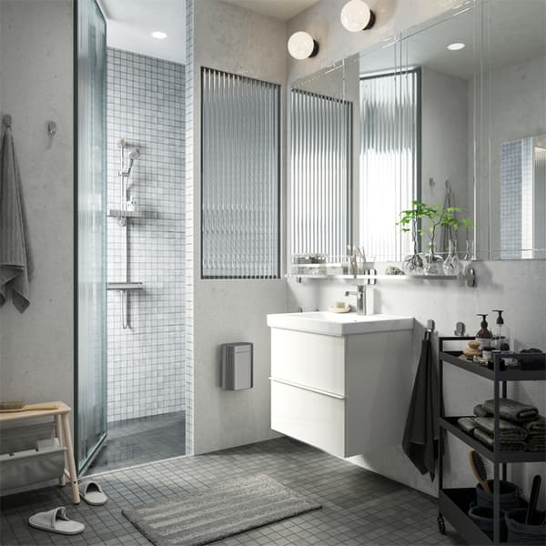 Ein in Weiß und Grau eingerichtetes Badezimmer mit Dusche, dem ODENSVIK Waschbecken und einem NISSAFORS Servierwagen in Schwarz für Handtücher, Seife und mehr