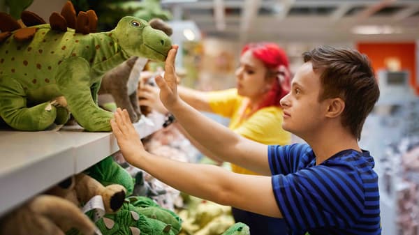 Ein junger Mann interagiert in der IKEA-Kinderabteilung mit einem grünen Plüschtier-Dinosaurier. Im Hintergrund sind weitere Stofftiere zu sehen. 