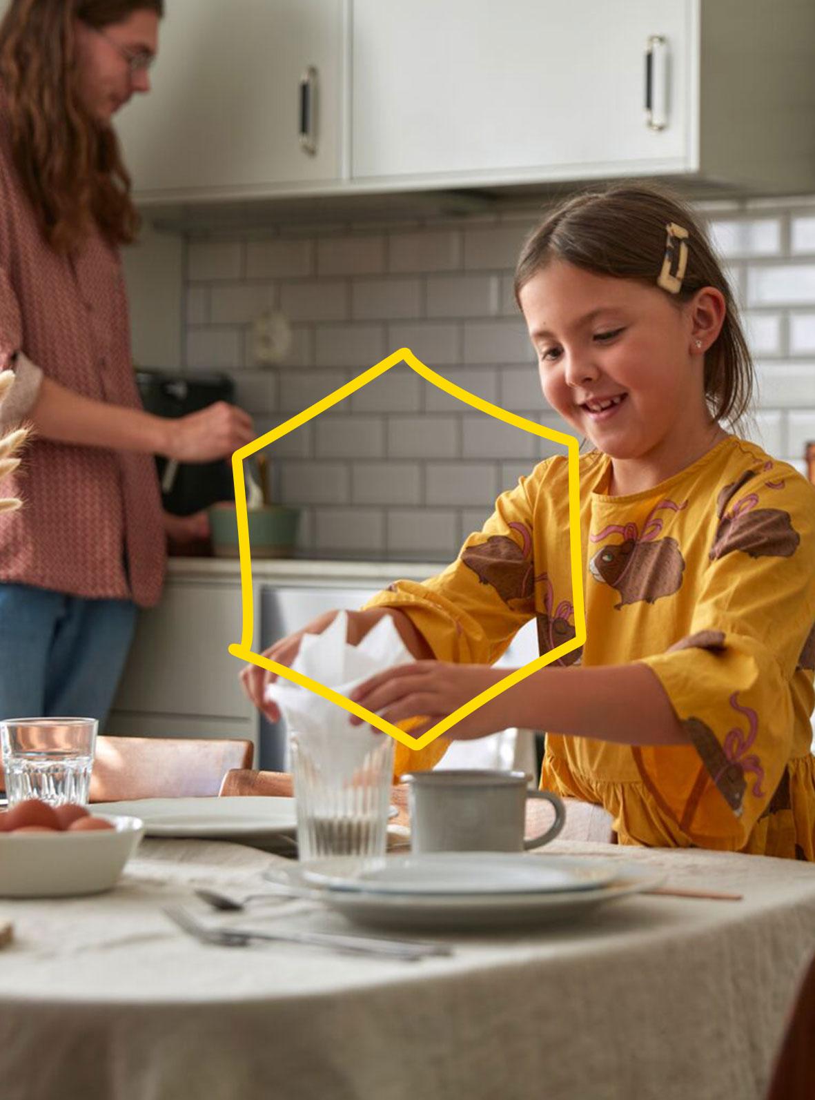 Ein Kind in einem gelben Kleid legt eine Serviette in ein Glas, während eine andere Person in einer hellen Küche mit weißen Fliesen Zutaten in einer Schüssel verrührt.