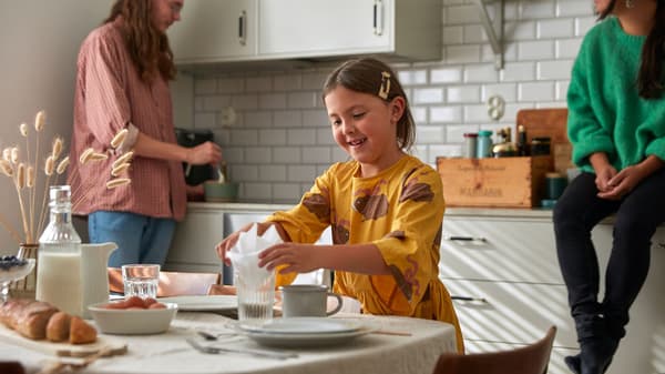 Ein Kind in einem gelben Kleid legt eine Serviette in ein Glas, während eine andere Person in einer hellen Küche mit weißen Fliesen Zutaten in einer Schüssel verrührt.
