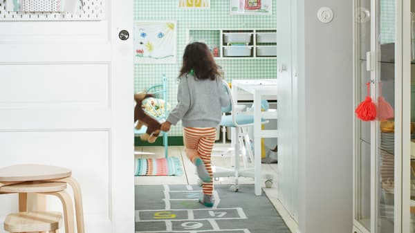 Ein Kind rennt durch eine offene Schiebetür eines in Weiß und Grün eingerichteten Zimmers mit Bett, Aufbewahrung, einem Schreibtisch und einem HEMMAHOS Teppich.
