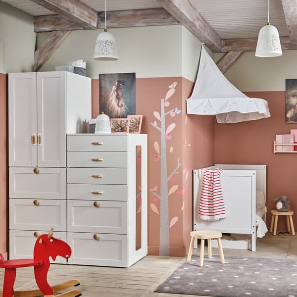 Ein Kinderzimmer in Weiß/Rosa mit einem Kleiderschrank und einem Babybett mit Betthimmel in Weiß im traditionellen Stil und einem roten Schaukelelch