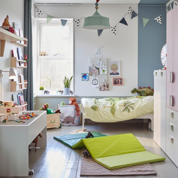Ein Kinderzimmer mit einem Kleiderschrank in Weiß/Blassrosa, einer grünen Gymnastikmatte, einem weißen Spieltisch mit Stauraum und Stofftierdinosauriern