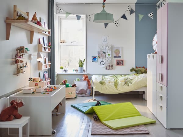Ein Kinderzimmer mit einem Kleiderschrank in Weiß/Blassrosa, einer grünen Gymnastikmatte, einem weißen Spieltisch mit Stauraum und Stofftier-Dinosaurier.